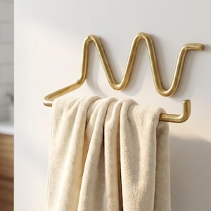 Pode incluir: Um toalheiro de metal dourado montado em uma parede branca. O toalheiro tem um design ondulado único e segura uma toalha bege. O ambiente do banheiro inclui um vislumbre de uma janela e um armário de madeira.