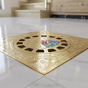 Könnte beinhalten: Ein quadratischer, goldfarbener Bodenablaufdeckel mit einem verzierten, gravierten Design. Die Mitte zeigt ein kreisförmiges Muster mit einem farbenfrohen Emblem. Der Ablaufdeckel ist in einen weißen Fliesenboden eingelassen.