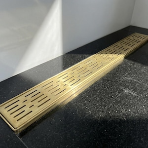 May include: A rectangular brass shower drain with a slotted design. The drain is installed on a black granite surface, with a white wall in the background. The drain is approximately 36 inches long.