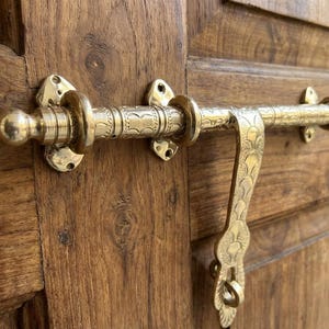May include: A brass door bolt with intricate etched designs, mounted on a wooden door. The bolt features a handle with a decorative pattern and a round knob. The door is made of wood with visible grain.