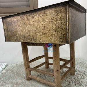 May include: A rectangular, hammered brass sink with a textured surface, mounted on a rustic wooden stool. The sink has a dark, aged appearance, and the stool features natural wood legs and supports. The overall aesthetic is vintage and handcrafted.