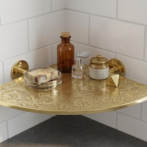 May include: A decorative brass corner shelf with an etched floral design, holding a soap dish, amber glass bottle, perfume bottle, small jar, and a gold geometric object. The shelf is mounted on a white tiled wall.