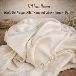 Pode incluir: Tecido creme com um padrão de tecido de diamante. O tecido está drapeado, mostrando dobras macias e uma borda com franjas. O texto "JPHandloom" e "100% Eri Vegan Silk Diamond Weave Pattern Fabric" são visíveis.