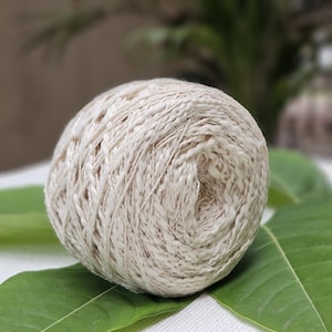 May include: A ball of off-white yarn with a subtle shimmer, resting on green leaves.
