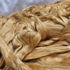 May include: Close-up of a pile of golden brown fibers, likely raw silk or a similar natural fiber. The fibers are soft and fluffy, with a slightly textured appearance.