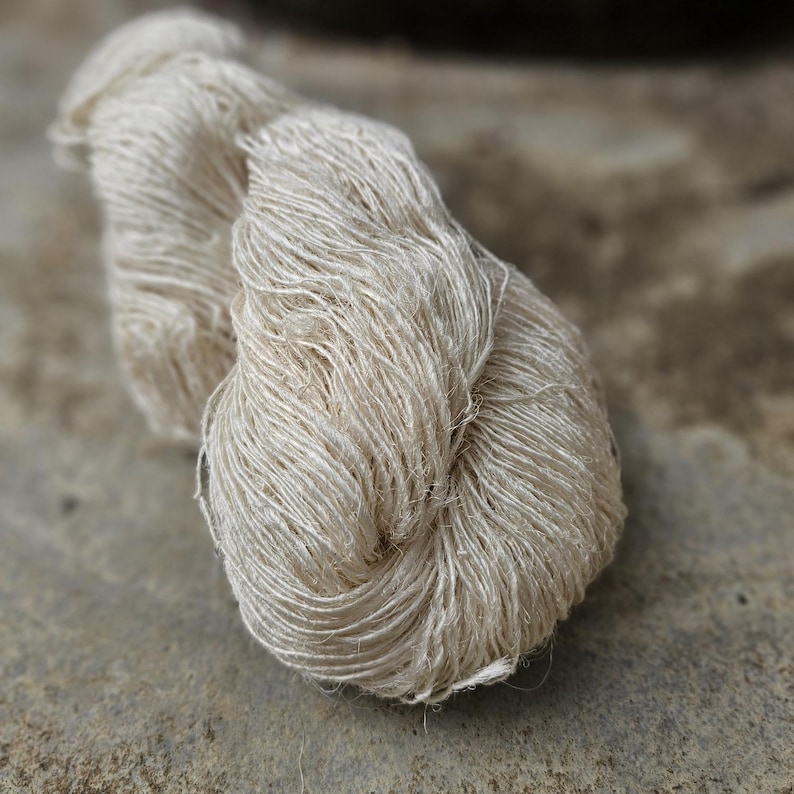 May include: A close-up shot of a skein of natural-colored yarn. The yarn appears to be made of a fine, silky fiber, with a soft, slightly textured appearance. The yarn is tightly wound, and the color is a pale, creamy beige.