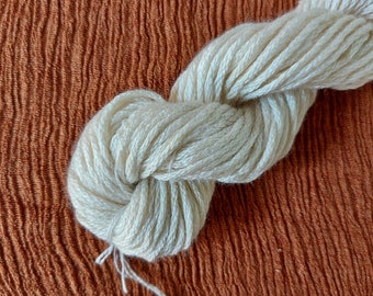 Peut inclure: Une pelote de fil blanc cassé est nouée et torsadée, avec un brin lâche qui s'étend du paquet. Le fil est posé sur un fond de tissu texturé de couleur rouille. Le fil semble doux et prêt pour le travail.