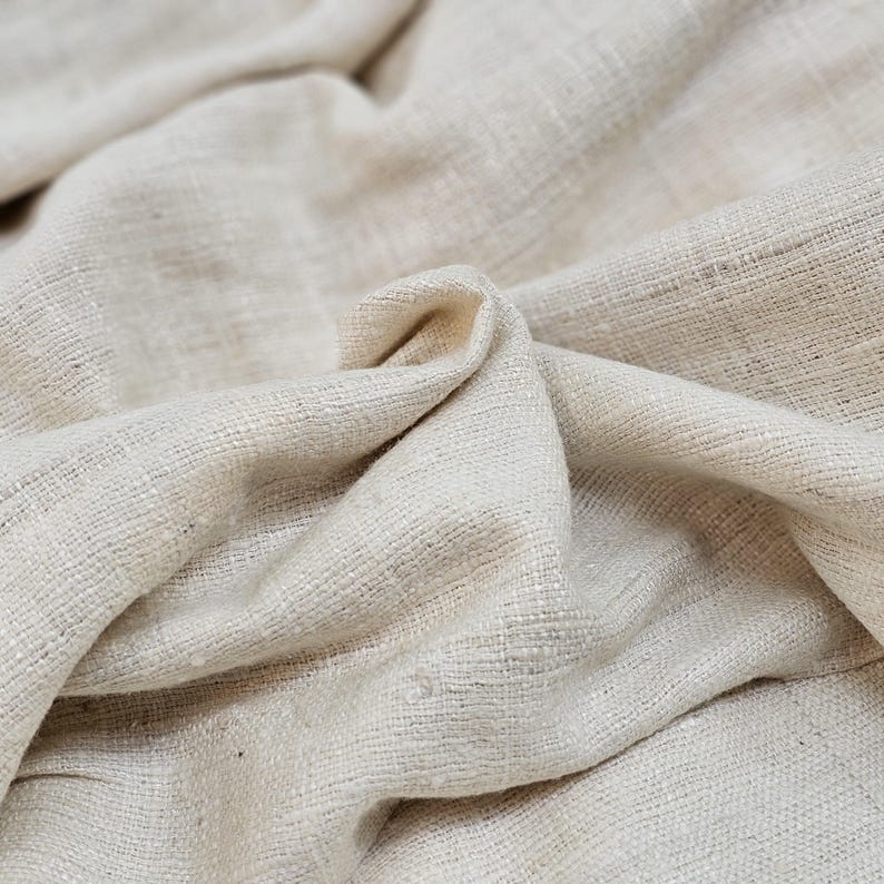May include: Close-up of a natural off-white fabric with a textured weave. The fabric is folded and draped, showing its soft and drapey texture.