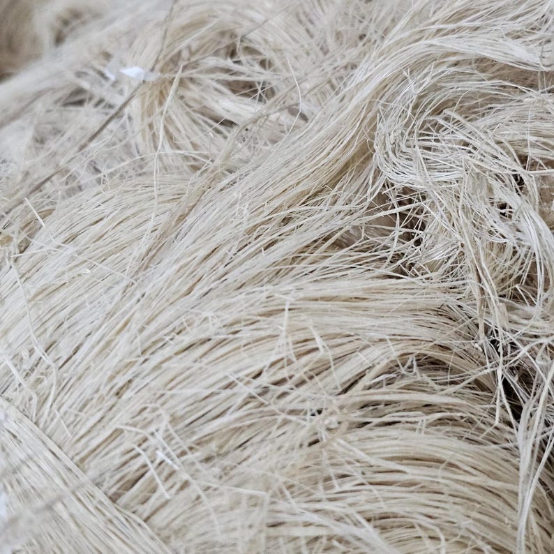 K&ouml;nnte beinhalten: Nahaufnahme eines B&uuml;ndels naturfarbener Fasern. Die Fasern sind lang, d&uuml;nn und haben eine leicht raue Textur. Die Fasern sind miteinander verflochten und scheinen lose miteinander verbunden zu sein, wodurch eine dichte, strukturierte Masse entsteht. Die Farbe ist hellbeige.