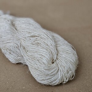 May include: A skein of off-white yarn, tightly wound and textured. The yarn appears to be made of natural fibers, with a slightly irregular, handmade appearance. The neutral color and texture suggest it could be used for knitting or other fiber crafts.