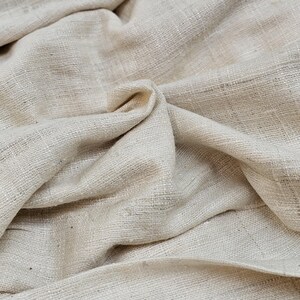 May include: A close-up of a natural, off-white fabric with a textured weave. The fabric is folded and draped, showing its soft and drapey texture.