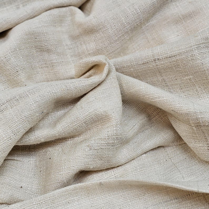 May include: A close-up of a natural, off-white fabric with a textured weave. The fabric is folded and draped, showing its soft and drapey texture.