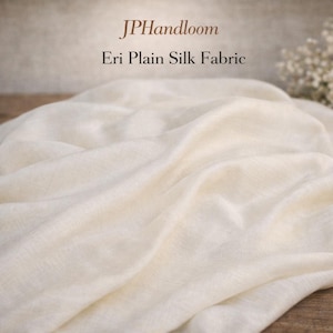 Puede incluir: Tela de seda Eri lisa de color crema, drapeada y texturizada, con el texto "JPHandloom" y "Eri Plain Silk Fabric" en marrón. La tela parece suave y fluida, con sutiles variaciones de tono.