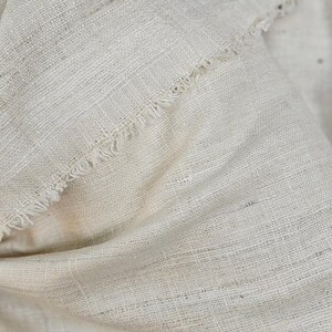 May include: A close-up of a natural off-white fabric with a textured weave. The fabric has a frayed edge.