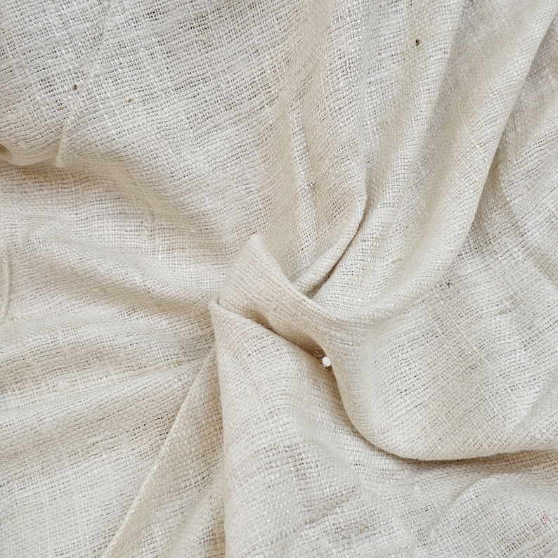 May include: Close-up of a natural off-white fabric with a woven texture. The fabric is folded and wrinkled, showing its soft and drapey quality.