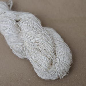 May include: A close-up of a skein of undyed, natural-colored yarn. The yarn is tightly twisted and has a slightly textured appearance. The neutral color and natural fibers suggest it is suitable for various knitting or crochet projects.