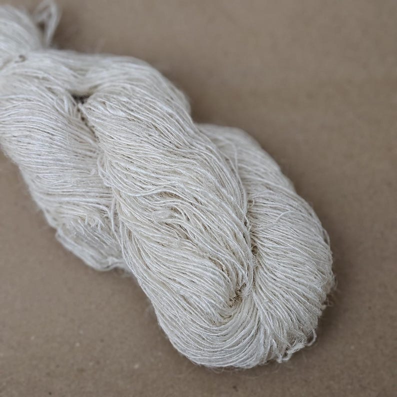 May include: A close-up of a skein of undyed, natural-colored yarn. The yarn is tightly twisted and has a slightly textured appearance. The neutral color and natural fibers suggest it is suitable for various knitting or crochet projects.