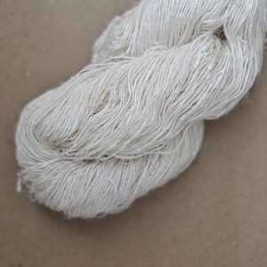 May include: A close-up of a skein of undyed, off-white yarn. The yarn is loosely spun, with visible fibers and a slightly textured appearance. The background is a solid, neutral brown.