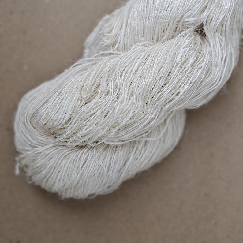 May include: A close-up of a skein of undyed, off-white yarn. The yarn is loosely spun, with visible fibers and a slightly textured appearance. The background is a solid, neutral brown.