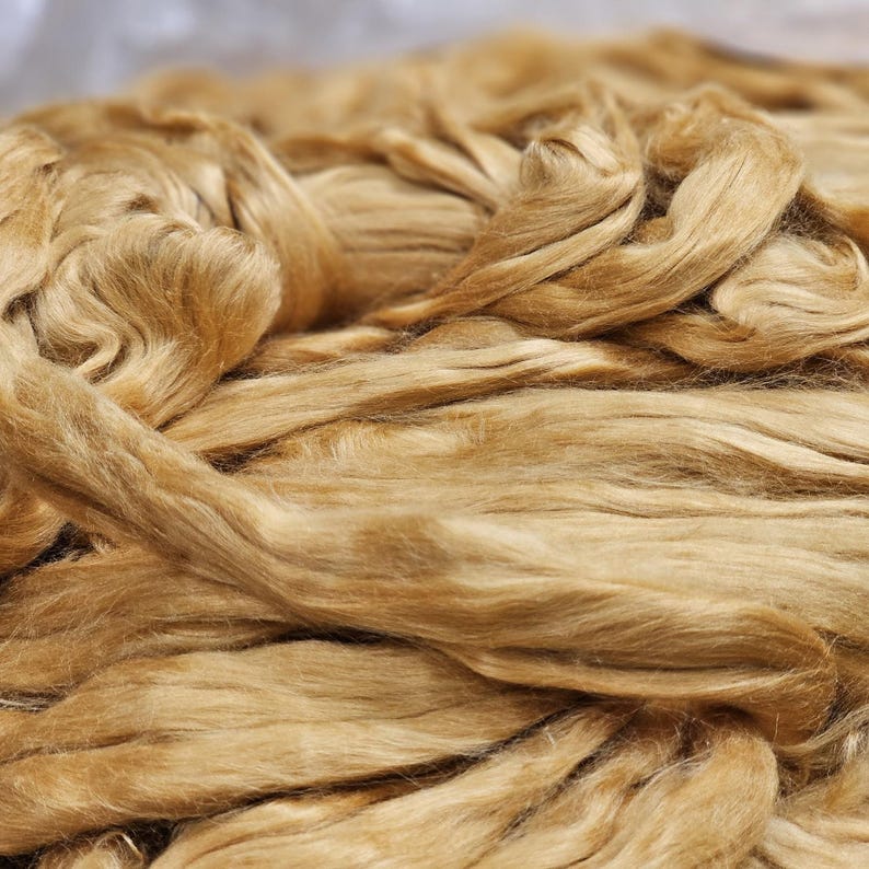 May include: Close-up of a pile of golden brown raw silk fibers. The fibers are soft and fluffy, and they are arranged in a random pattern.