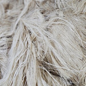 K&ouml;nnte beinhalten: Nahaufnahme eines Haufens nat&uuml;rlicher, ungef&auml;rbter Flachsfasern. Die Fasern sind lang, d&uuml;nn und haben eine hellbeige Farbe. Die Textur wirkt weich und leicht verknittert, was auf einen rohen, unverarbeiteten Zustand hindeutet.
