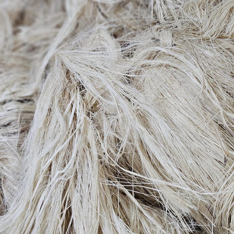 K&ouml;nnte beinhalten: Nahaufnahme eines Haufens nat&uuml;rlicher, ungef&auml;rbter Flachsfasern. Die Fasern sind lang, d&uuml;nn und haben eine hellbeige Farbe. Die Textur wirkt weich und leicht verknittert, was auf einen rohen, unverarbeiteten Zustand hindeutet.