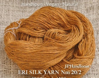 Peut inclure: Pelote de fil de soie Eri, de couleur brun doré. Le texte indique "ERI SILK PEACE SILK Handspun Vegan Natural" et "ERI SILK YARN Nm 20/2 ~1000 Yards / 100 g". Le fil est filé à la main et végétalien, environ 914 mètres pour 100 grammes.