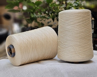 May include: Two spools of natural colored yarn, one on top of the other, on a white fabric surface.