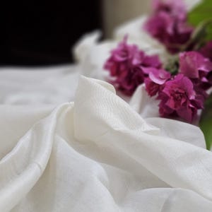 Puede incluir: Tela de seda blanca con una textura suave y fluida. La tela está drapeada sobre una superficie y tiene un brillo sutil. Se pueden ver flores rosas en el fondo.