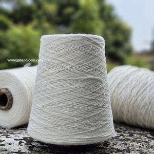 Puede incluir: Tres carretes de hilo de algodón blanco, enrollados apretados y listos para tejer o hacer punto. El hilo es natural y sin blanquear, con una superficie suave y ligeramente texturizada.