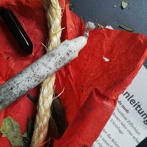 Può includere: Una candela bianca con fuliggine nera, una corda, un cristallo nero e foglie verdi su un pezzo di carta velina rossa. La carta ha un pezzo di carta bianca sopra con il testo "Anleitung L" e del testo tedesco.
