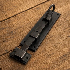 Puede incluir: Un cerrojo de puerta de metal negro que presenta una placa de montaje rectangular con orificios pretaladrados, un mecanismo de barra deslizante y un asa circular en la parte superior. El herraje tiene un acabado mate oscuro y un diseño robusto.