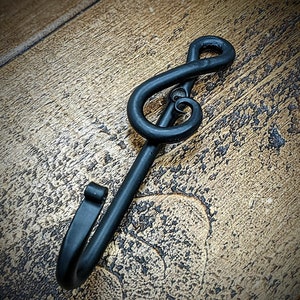 Könnte beinhalten: Ein schwarzer, handgeschmiedeter Metallhaken in Form eines Notenschlüssels. Der Haken besteht aus massivem Metall und hat eine matte Oberfläche. Das Design zeigt eine klassische Notenschlüsselform mit einem gebogenen Haken am unteren Ende. Der Haken ist zum Aufhängen von Gegenständen bestimmt.