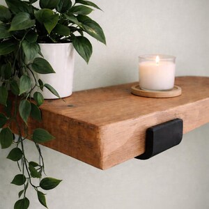May include: A wooden shelf with a lit candle in a glass jar and a small plant in a white pot. The shelf is supported by a black metal bracket. The plant has green leaves.