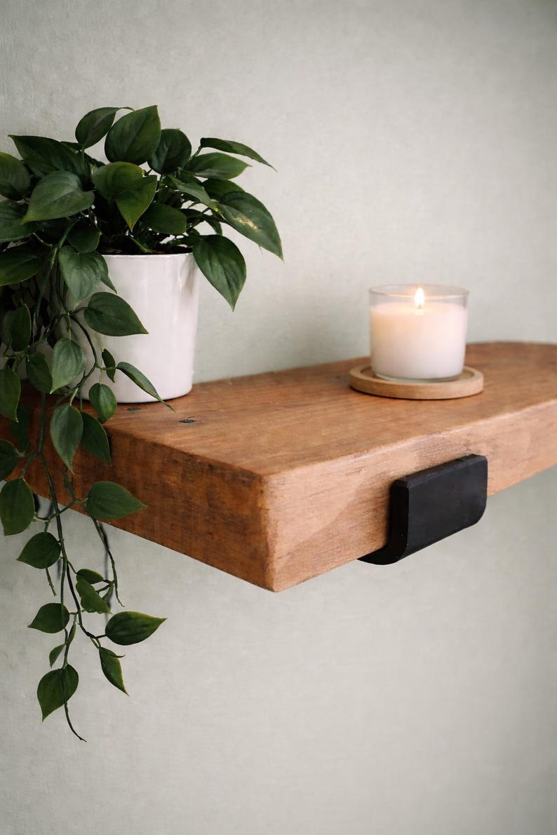 May include: A wooden shelf with a lit candle in a glass jar and a small plant in a white pot. The shelf is supported by a black metal bracket. The plant has green leaves.