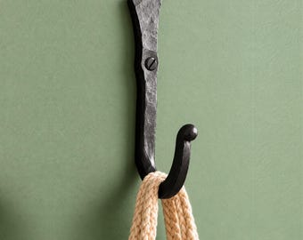 Könnte beinhalten: Ein schwarzer Wandhaken aus Metall mit gehämmertem, texturiertem Finish und einer einzelnen Befestigungsschraube. Der Haken hat einen gebogenen unteren Arm und einen flachen, länglichen oberen Teil. Ein dickes, beiges geflochtenes Seil hängt am Haken.