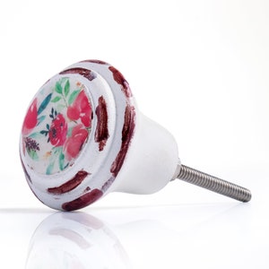 May include: White ceramic cabinet knob with a floral design. The knob features a painted floral pattern in shades of red and green on a white background. The edges are distressed with brown paint, and a metal screw extends from the back.