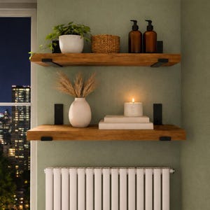 May include: Two wooden floating shelves with black metal brackets are mounted on a green wall. The top shelf holds a potted plant, a woven basket, and two brown bottles. The bottom shelf displays a vase, books, and a lit candle.