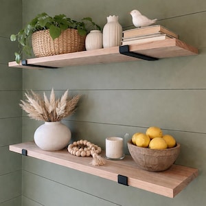 May include: Two wooden floating shelves with black metal brackets are mounted on a green wall. The top shelf holds a woven basket with a plant, vases, and a bird figurine. The bottom shelf displays a vase with dried grass, a wooden bowl of lemons, and decorative beads.