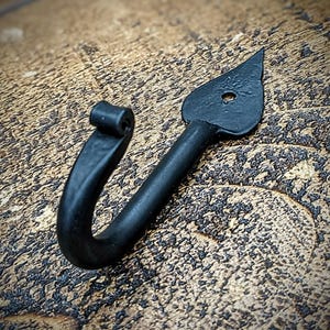 May include: Black metal hook with a pointed tip and a rounded loop. The hook is designed for hanging items on a wall.