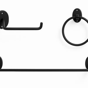 May include: A set of three matte black bathroom fixtures: a toilet roll holder, a towel ring, and a towel rail. Each piece features an oval mounting plate and a simple, functional design. The fixtures are mounted on a white wall.