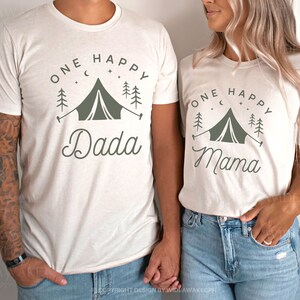 Op de afbeelding: Twee witte T-shirts met een kampeerthema. De shirts hebben een groene tent, bomen en sterren. E&eacute;n shirt zegt "One Happy Dada" en de andere zegt "One Happy Mama".