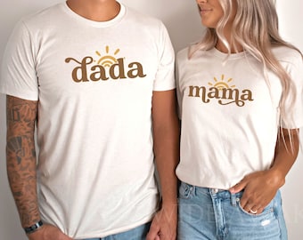 Peut inclure: Deux t-shirts blancs avec un soleil et les mots "dada" et "mama" en texte brun.