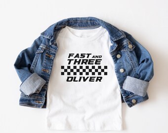 May include: White t-shirt with black text that reads "Fast and Three Oliver" with a black and white checkered pattern underneath.