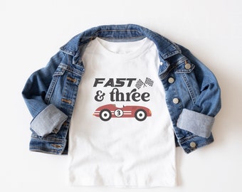 May include: White t-shirt with a red race car graphic and the text "Fast & three" in black. The shirt is on a white surface with a blue denim jacket folded on top.