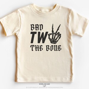 Puede incluir: Una camiseta de color crema con texto negro que dice "Bad Two The Bone" y una mano de esqueleto haciendo un signo de paz.