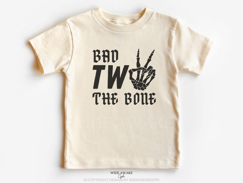 Puede incluir: Una camiseta de color crema con texto negro que dice "Bad Two The Bone" y una mano de esqueleto haciendo un signo de paz.