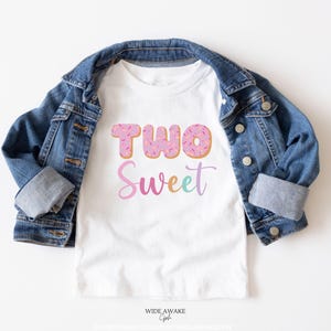May include: White t-shirt with the text "TWO Sweet" in pink, blue, and purple. The "TWO" is made up of two pink doughnuts with colourful sprinkles.