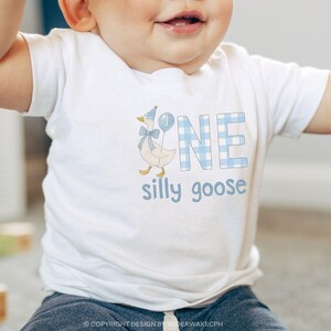 K&ouml;nnte beinhalten: Ein wei&szlig;es Kurzarm-T-Shirt mit einer Grafik einer Gans, die einen blauen Partyhut und eine Fliege tr&auml;gt, neben einem blauen Luftballon mit der Zahl eins. Das Wort ONE ist in blauem Karomuster gedruckt, darunter steht der Text silly goose.