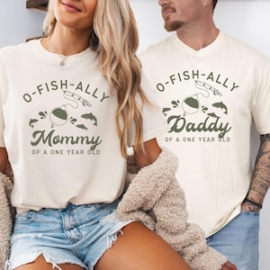 Pu&ograve; includere: T-shirt color avorio con grafiche coordinate. Una maglietta dice "Mommy of a One Year Old" e l'altra "Daddy of a One Year Old". Il design include un galleggiante da pesca, una canna da pesca e dei pesci.
